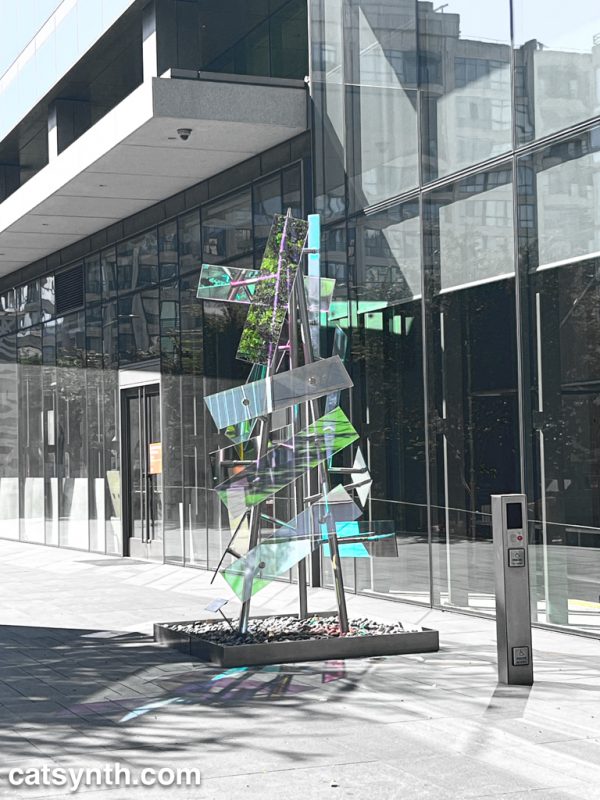 Sculpture made of steel and glass with brightly colored reflections.  The steel and glass facade and ground are desaturated, bringing out the color reflections in the sculpture as well as the light passing through it to the ground.