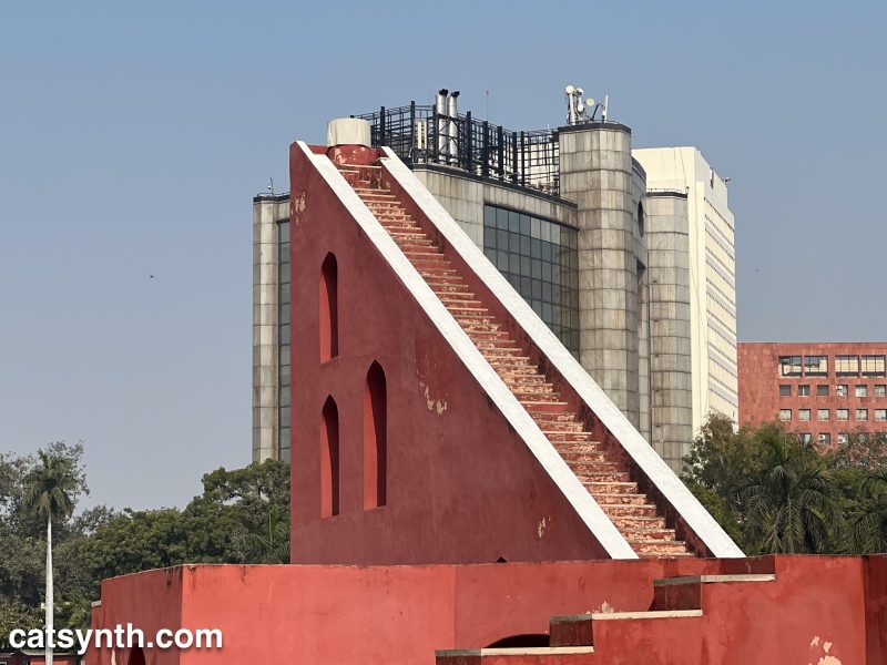 The triangular center tower of the Samrat Yantra at Juntar Muntar in New Deli.  There is a large modern building behind it.