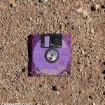 Purple translucent 3.5-inch floppy disk sitting on a flat patch of desert ground with sand and pebbles.