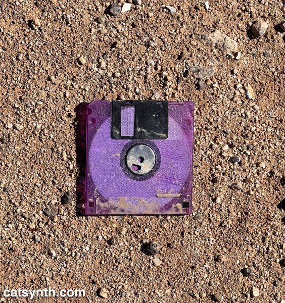 Purple translucent 3.5-inch floppy disk sitting on a flat patch of desert ground with sand and pebbles.
