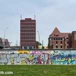 Surviving section of the Berlin Wall covered in grafiti. Behind it we see some modern and traditional buildings, and the corner of a large train station.