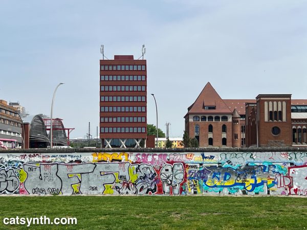 Surviving section of the Berlin Wall covered in grafiti. Behind it we see some modern and traditional buildings, and the corner of a large train station.