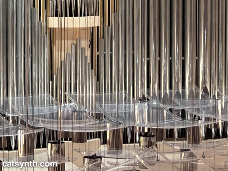 Upper pipes of a large pipe organ, seen above plexiglass curved ceiling plates and crome lighting fixtures.