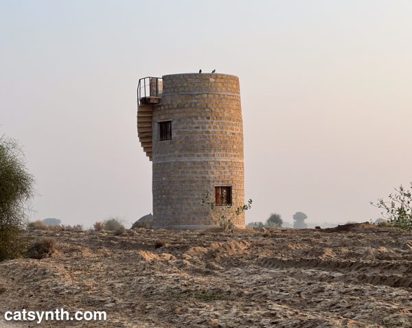 Small cylindrical two-story tower made of sandstone with a spiral staircase on the outside, sitting in a desert landscape.