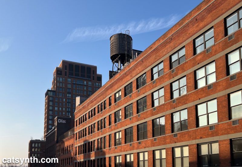 A long red brick building in perspective, in New York with a water tower on top.  Behind it is a taller, newer apartment building.  All the buildings are set against a blue sky and late afternoon winter sunlight.