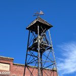 Bell tower with a weathervane in front of an brick building with the sign "Masonic Temple 1893". The sky is a brilliant blue and has a few wispy cirrus clouds.