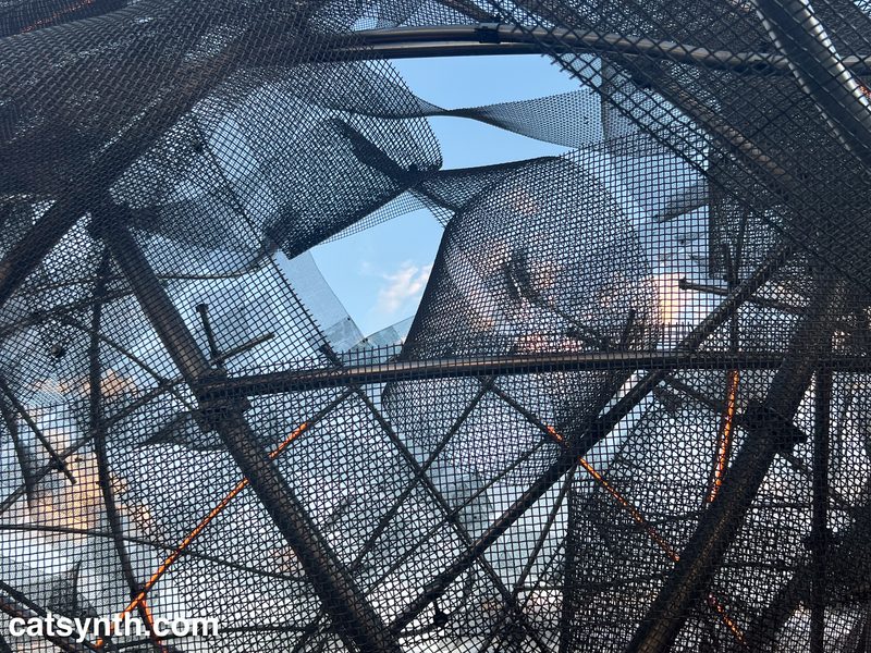 Looking up at a blue sky through curved mesh and metal beam formations.
