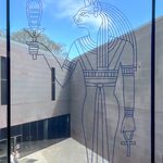 Outline of Egyptian cat goddess Bastet on a window overlooking an angular ultra-modernist courtyard. There is a bright blue sky in the background with wispy clouds.