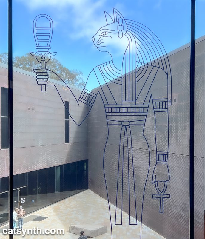 Outline of Egyptian cat goddess Bastet on a window overlooking an angular ultra-modernist courtyard.  There is a bright blue sky in the background with wispy clouds.