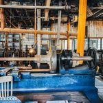 Old metalworking machinery in a large shop space with lots of gears, wheels, axels, and rusting pipes. There is an old chair in the foreground.