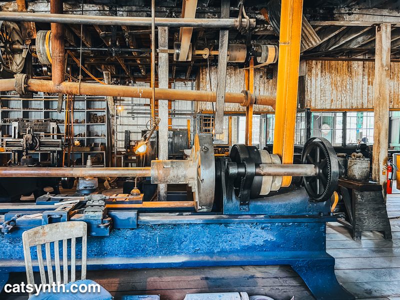 Old metalworking machinery in a large shop space with lots of gears, wheels, axels, and rusting pipes.
There is an old chair in the foreground.
