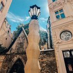Looking upwards near the base of the zig-zagging structure of the cubist lamp post in Prague. We see a blue sky with wispy clouds and older buildings above and to the sides, respectively.