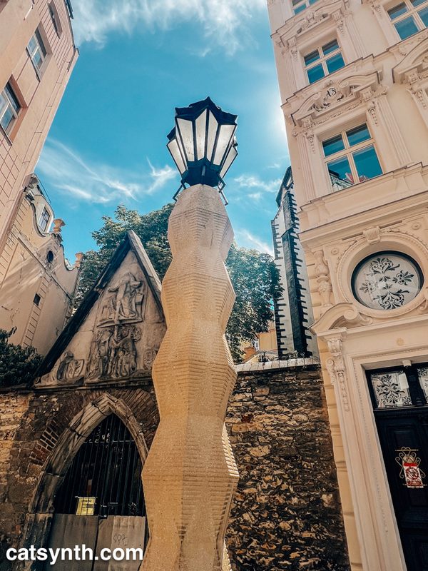 Looking upwards near the base of the zig-zagging structure of the cubist lamp post in Prague.  We see a blue sky with wispy clouds and older buildings above and to the sides, respectively.