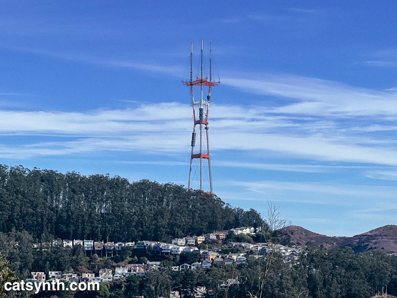 Sutro Tower (giant communications antenna) in San Francisco above a hill covered with trees and crisscrossed with houses.